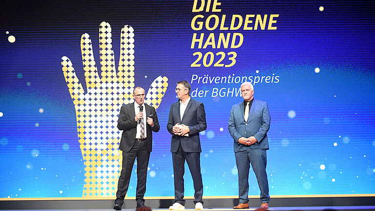 Drei Männer stehen auf der Bühne, im Hintergrund ist die GOLDENE Hand zu sehen: Die Vorstandsvorsitzenden Roland Kraemer (links) und Manfred Wirsch (rechts) im Gespräch mit Moderator Karl-Josef Thielen. 