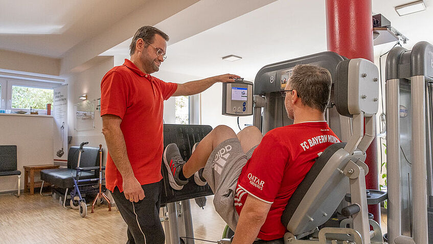 Ein Trainingsraum mit Fitnessgeräten: In der Mitte des Bildes steht Simon Emmel, Physiotherapeut bei Steven Wedhorn, der rechts an der Beinpresse sitzt. Emmel schaut auf ein Display. .