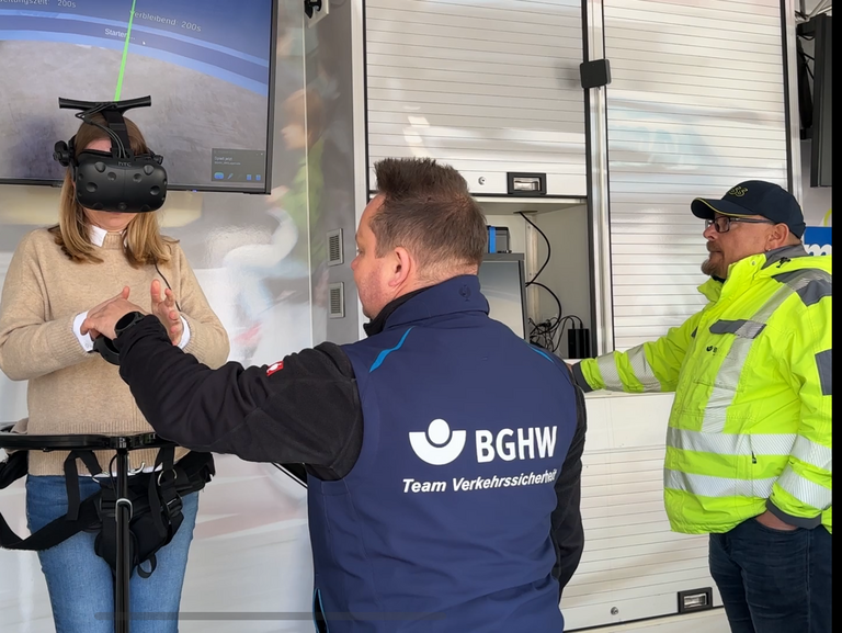  Eine Frau steht im BGHW mobil in einem Stahlgestell, ein sogenannter Virtualizer. Sie hat eine VR-Brille an, die sie in die Virtuelle Welt eines Materiallagers eintauchen lässt. Rechts von ihr stehen zwei Männer. Die BGHW-Trainer erklären ihr, wie die Technik funktioniert. 