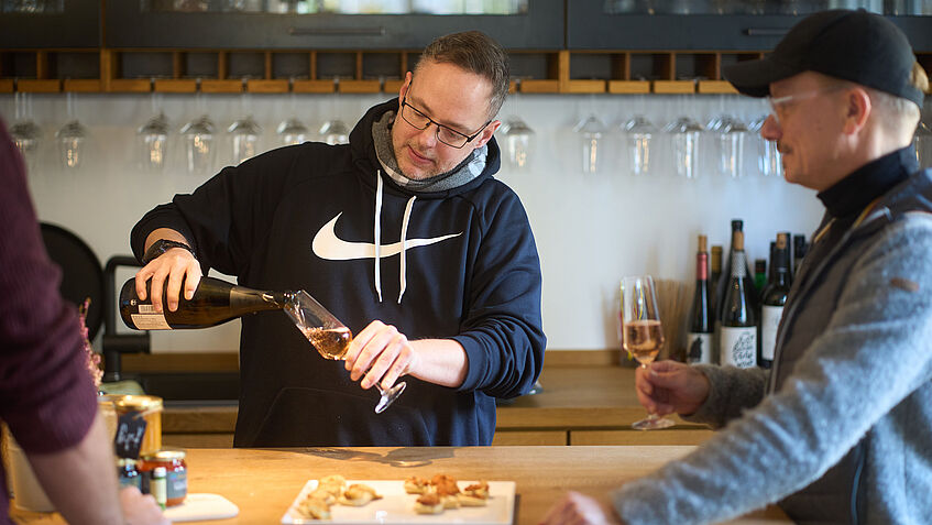 Steven Wedhorn im schwarzen Sweatshirt steht hinter einer Theke und schenkt Wein in ein Glas, hinter ihm ist ein Regal mit Gläsern zu sehen. Rechts steht ein Mann und schaut ihm zu, links ist die Hand eines weiteren Gastes zu sehen..