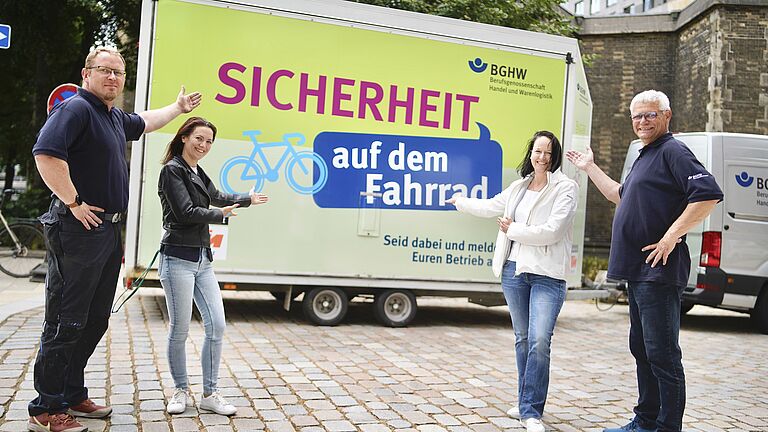 Vor einem hellgrünen Pkw-Anhänger mit dem Logo der BGHE, der Aufschrift "Sicherheit auf dem Fahrrad" und einem aufgemalten Fahrrad stehen zwei Frauen und zwei Männer. Sie zeigen auf den Schriftzug. 
