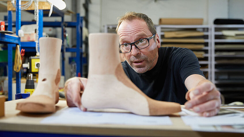 Ein Mann im schwarzen T-Shirt sitzt in einer Werkstatt und betrachtet zwei Holz-Leisten. Es sind die Muster, nach denen der Orthopädie-Schuhmachermeister die Maßschuhe fertigt. 