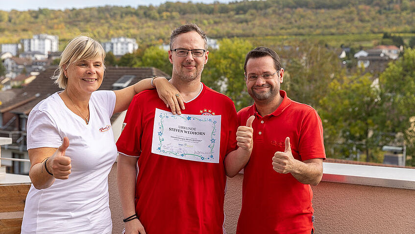 (Von links) Christine Blass, Steven Wedhorn und Simon Emmel stehen auf einem Dachbalkon und halten lachend eine Urkunde in die Kamera. Sie ist eine Anerkennung für Stevens erfolgreiche Brainrestart-Intensivtherapie.