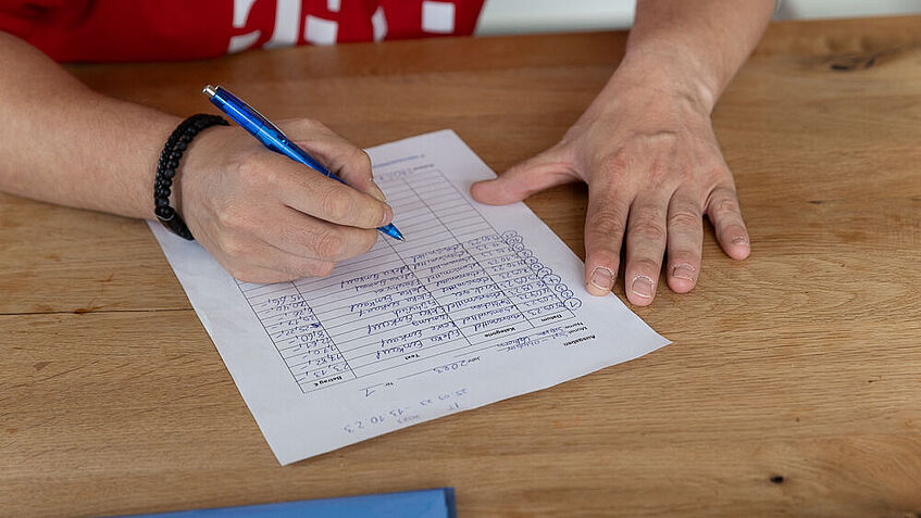 Auf einem DIN-A-4-Blatt, das auf einem Holztisch liegt, erstellt eine Person handschriftlich mit einem blauen Filzstift eine Einkaufsliste. Man sieht nur die Hände und Unterarme der Person und den Ausschnitt eines roten Trikots. 