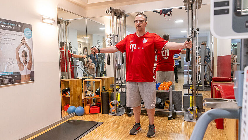 Steven Wedhorn trainiert beim Armtraining im Fitnessraum. Er hat die Arme ausgebreitet und zieht an beiden Seiten an Seilzügen. 