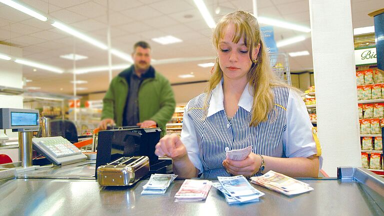 Eine Kassiererin zählt Geld im Supermarkt, ein Kunde nähert sich von hinten