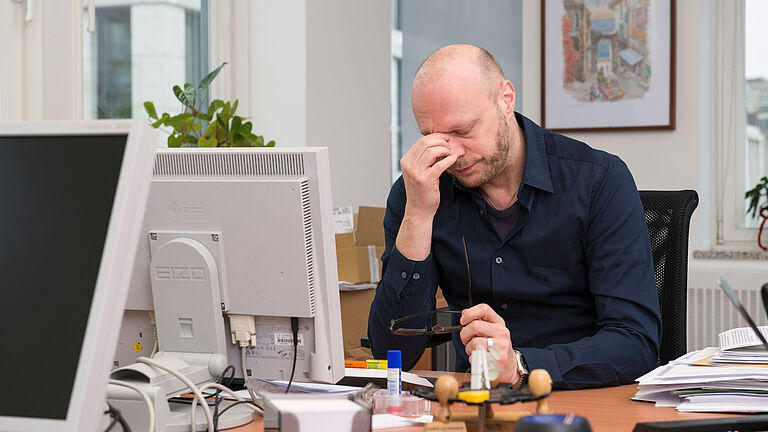 Ein Mann im blauen Hemd sitzt mit geschlossenen Augen vor einem Bildschirm am Schreibtisch und zieht mit der linken Hand die Augenbrauen zusammen. Er wirkt müde.