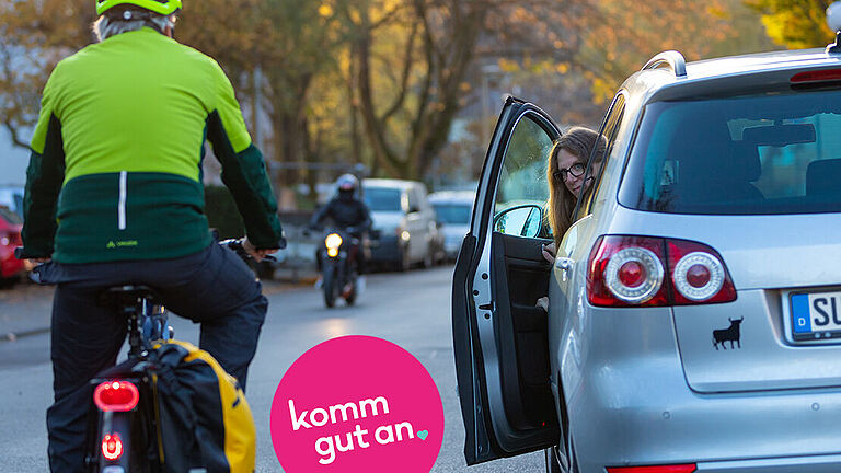 Eine Person auf einem Fahrrad mit gelbem Helm und grüner Jacke fährt auf einer Straße, während eine Frau in einem silbernen Auto mit der rechten Hand vorsichtig die Fahrertür öffnet und sich nach dem Radfahrer umschaut. Ein Motorradfahrer nähert sich im Hintergrund. Die Szene spielt sich in einer herbstlichen Umgebung mit Laubbäumen ab. Ein pinker Kreis mit der Aufschrift 'komm gut an' ist im Vordergrund sichtbar.