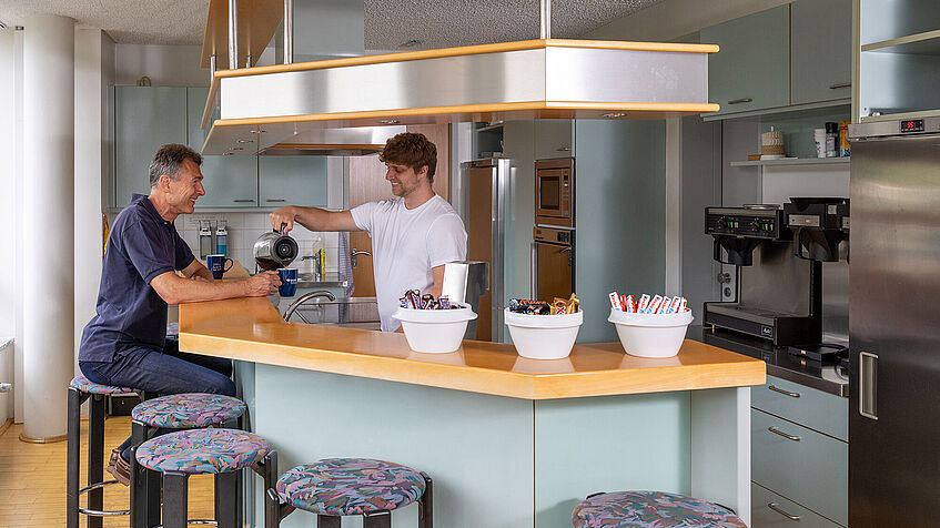 Küche mit Tresen. Ein Mann sitzt auf einem Barhocker. Ein anderer Mann steht hinter dem Tresen und schüttet ihm Kaffee in eine Tasse. Küche mit Tresen. Ein Mann sitzt auf einem Barhocker. Ein anderer Mann steht hinter dem Tresen und schüttet ihm Kaffee in eine Tasse.