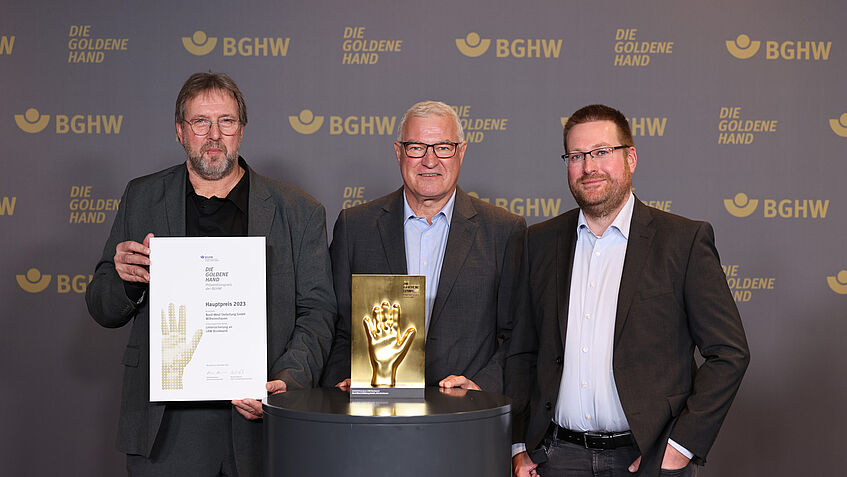 Gut gelaunt stehen drei Männer, Beschäftigte des Preisträgers Nord-West Oelleitung vor der BGHW-Fotowand mit dem Schriftzug GOLDENE HAND und zeigen die Goldene Hand und eine Urkunde in die Kamera.