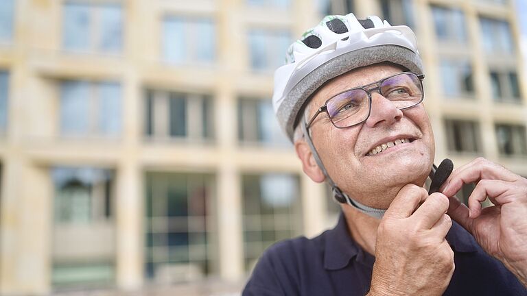 Das Bild zeigt den vorderen Rahmen eines schwarzen Pedelecss. An jeder Seite des Lenkers hängt ein Helm, links ein grüner, an der rechten Seite ein grauer Helm. Im Hintergrund ist eine mehrstöckige Hausfassade zu sehen.