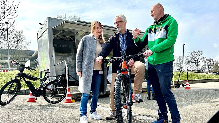 Claudia Kilian und Karl-Heinz Spiegel (Mitte) stehen zusammen mit E-Bike-Aussteller Frank Schnabel. Spiegel, der Mann in der Mitte sitzt auf einem E-Bike. Im Hintergrund steht das „BGHW mobil“.