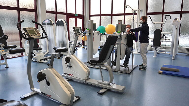 In einem Therapieraum der UBS Berlin stehen viele Trainingsgeräte im Vordergurnd. Hinten sitzt an ein Mann an einem Fitnessgerät, eine Therapeutin steht neben ihm und erklärt ihm das Gerät. 