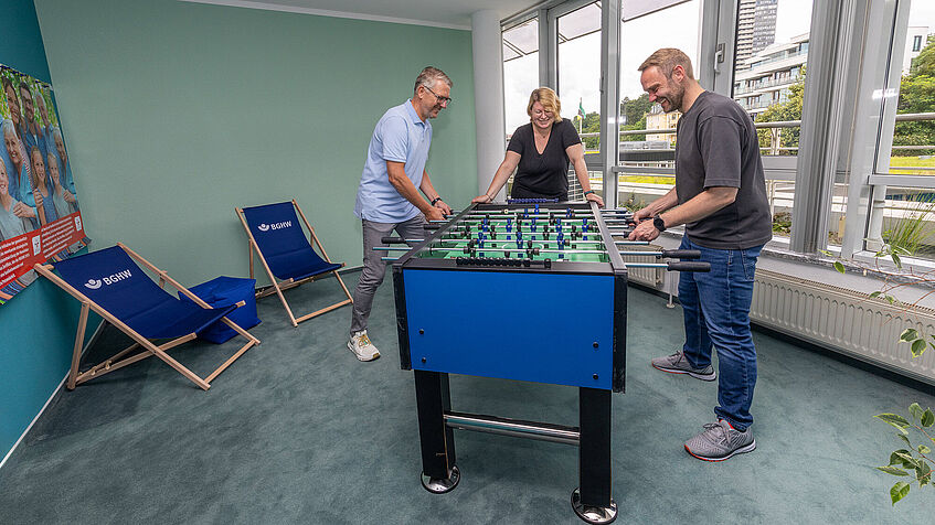 Zwei Männer spielen an einem Tisch Fußball Gerät. Eine Frau steht dabei und schaut zu. Zwei Männer spielen an einem Tisch Fußball Gerät. Eine Frau steht dabei und schaut zu.
