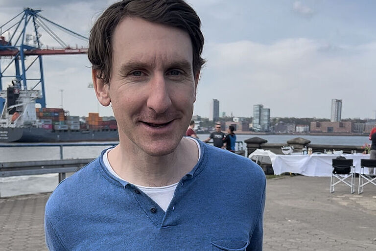 Thomas Springer steht mit seinem Fahrrad am Hafen in Hamburg und lächelt in die Kamera.