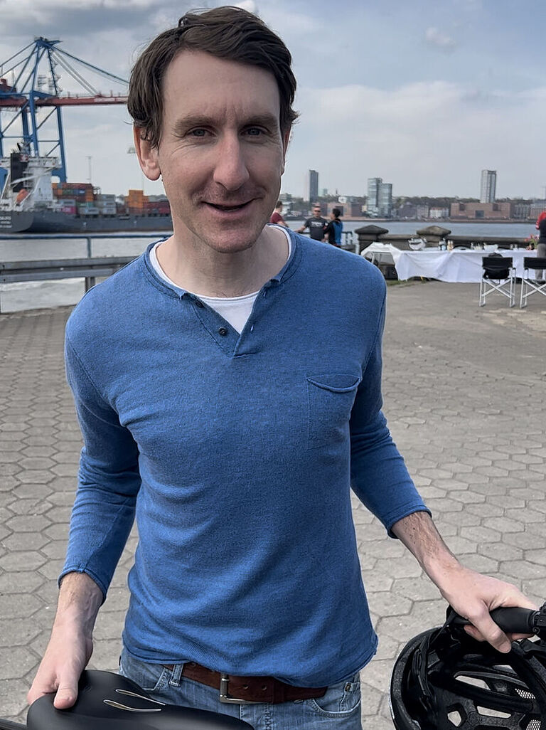 Thomas Springer steht mit seinem Fahrrad am Hafen in Hamburg und lächelt in die Kamera.
