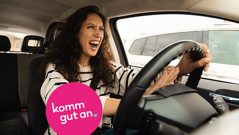 Eine junge Frau mit langen dunklen Haaren sitzt am Steuer eines Autos. Sie wirkt wütend, hat den Mund weit geöffnet, als würde sie schreien, und drückt mit beiden Händen auf die Hupe. Im Hintergrund sind andere Fahrzeuge zu sehen. In der linken unteren Bildecke befindet sich ein auffälliger pinker Kreis mit weißer Schrift, auf dem steht: „komm gut an“. Das Bild vermittelt eine angespannte Situation im Straßenverkehr.