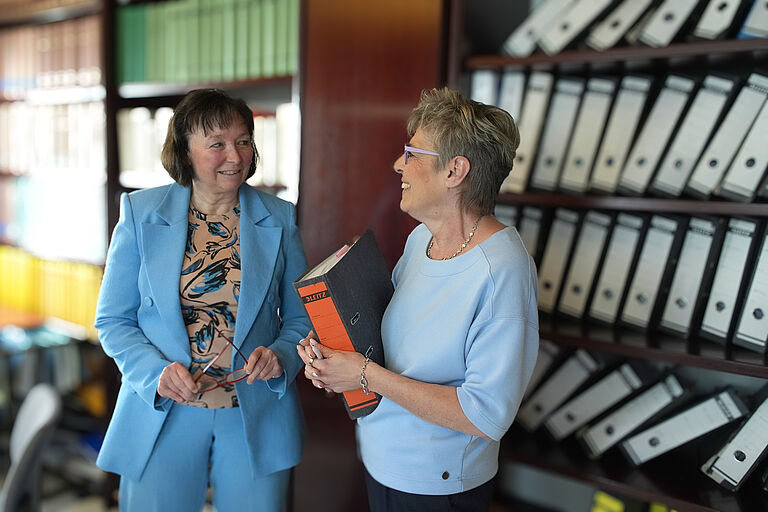 Ute Grötzinger (rechts) und Rechtsanwältin Sylvia Grasser stehen sich vor einem Regal voller Akten gegenüber und sprechen miteinander. Sie lachen sich an.