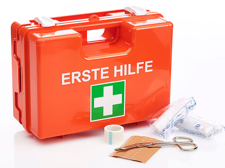 Ein orangener Erste-Hilfe-Verbandkasten, davor Schere, Pflaster und steriles Verbandmatarial