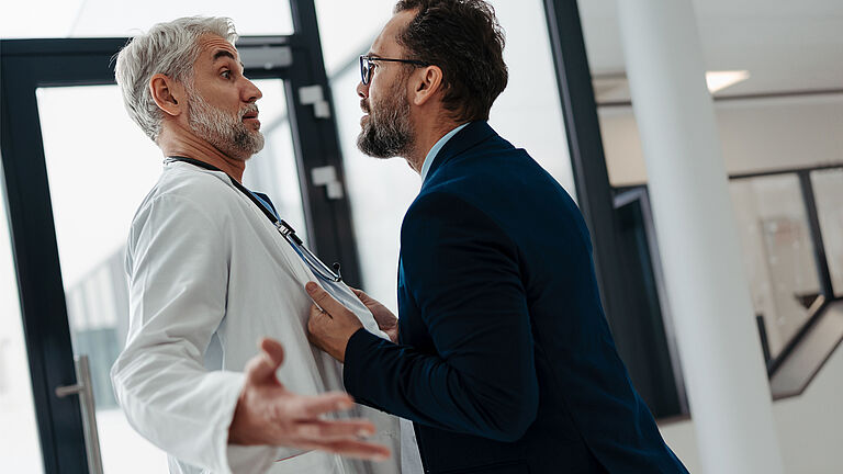 Ein Arzt im weißen Kittel und ein Mann, ein Patient in schwarzer Jacke stehen sich gegenüber. Der Mann zerrt am Kittel des Arztes und schaut ihn wütend an. Der Arzt, der ein Stethoskop um den Hals trägt, beugt sich nach hinten, um ihn auszuweichen.