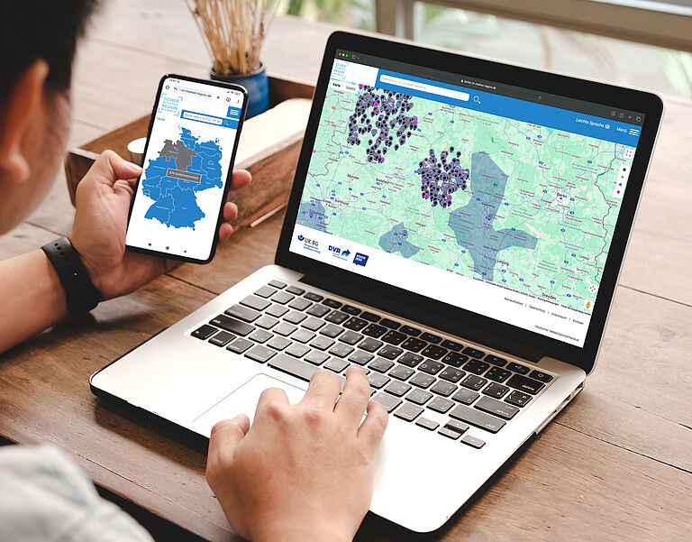 Junger Mann sitzt am Laptop und arbeitet mit der Anwendung "Sicher in meiner Region". Er hält ein Handy in der Hand. Die entsprechende App ist geöffnet.