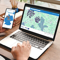 Sicher in meiner Region Junger Mann sitzt am Laptop und arbeitet mit der Anwendung "Sicher in meiner Region". Er hält ein Handy in der Hand. Die entsprechende App ist geöffnet.