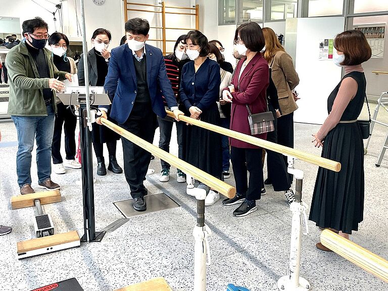 Eine Besuchergruppe aus Korea informiert sich in der BG-Klinik Ludwigshafen. Dabei schauen sie sich auch ein Gerät an, das ähnlich wie ein Stufenbarren ist 