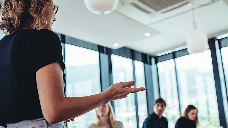 Eine Frau spricht in einem Seminar vor einer Gruppe Menschen, die um einen Tisch sitzen