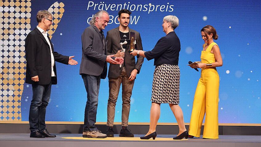 Blick auf die Bühne zur Präventionspreisverleihung: Links stehen drei Männer der Witt-Gruppe, die stolz die GOLDENE HAND von Silke Mayer-Seidler, Vorsitzende der Vertreterversammlung, entgegennehmen. Rechts im Bild steht Tina Babbel.