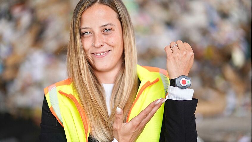 Standortleiterin Nina Vedder hält den linken Arm hoch und zeigt mit ihrer rechten Hand auf die Transponderuhr am linken Handgelenk. Im Hintergrund türmt sich das lose Altpapier.