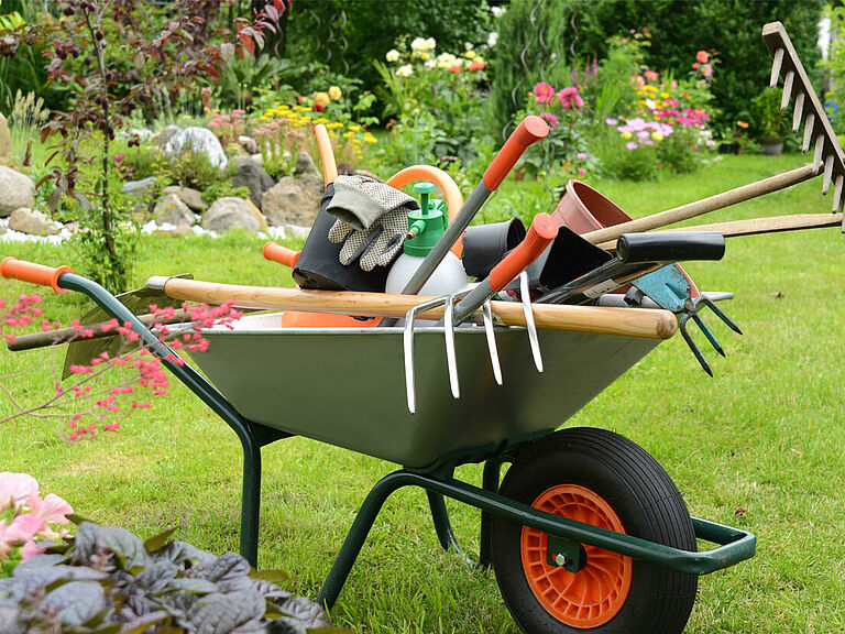 Schubkarren steht auf dem Rasen. Beladen mit Gartenwerkzeugen (wie Rechen, Spaten, Gartenhandschuhen etc.), die unordentlich übereinander liegen. 