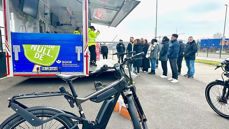Ein Mann steht mit einem Pedelec vor einer Gruppe von Menschen. Links von der Gruppe ist das BGHW-"mobil", ein Anhänger mit Display, Helmen und einem Laufband.