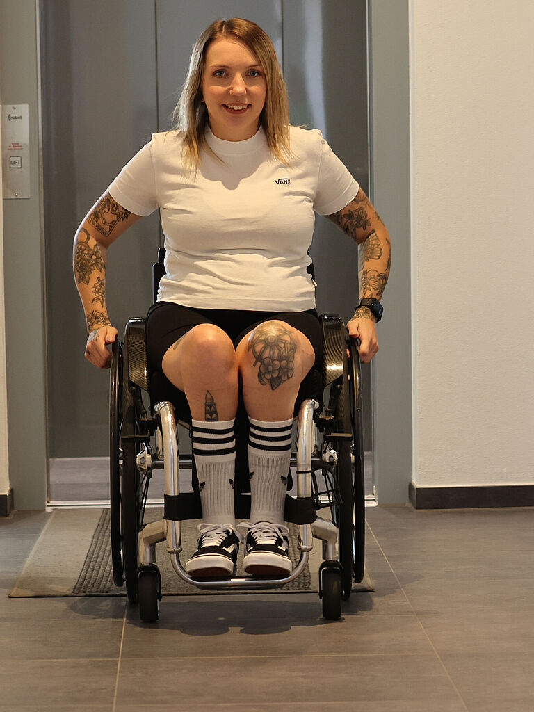 Tamara Fischer fährt mit ihrem Rollstuhl im Hausflur ihres Eigenheims in Richtung Kamera. Im Hintergrund ist ein Aufzug zu sehen, der mithilfe der BGHW eingebaut wurde. 
