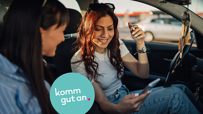 Zwei Personen sitzen in einem geparkten Auto. Die Person auf dem Fahrersitz hält ein Smartphone in der Hand und unterhält sich mit der Beifahrerin. Auf der Bildfläche ist zudem ein rundes Logo mit dem Schriftzug ‚komm gut an‘ zu sehen.