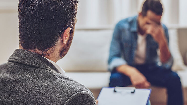 Zwei Männer in privater Atmosphäre im Gespräch. Sie sitzen sich gegenüber, ein Mann hat ein Klemmbrett in der Hand und notiert etwas. Der andere Mann wirkt bedrückt, hat den Kopf geneigt.