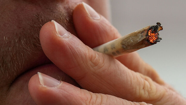  Eine Person raucht einen Joint, der zwischen Zeige- und Mittelfinger steckt. Man sieht nur einen Ausschnitt des Gesichtes, das durch die linke Hand der Person verdeckt ist, die an dem glühenden Joint zieht.