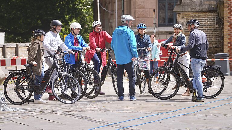 Eine Gruppe Frauen und Männer stehen mit ihren Pedelecs zusammen. Alle tragen Fahrradhelme, alle schauen den Mann in der blauen Jacke an. Er ist der Fahrtrainer und gibt wichtige Informationen. 