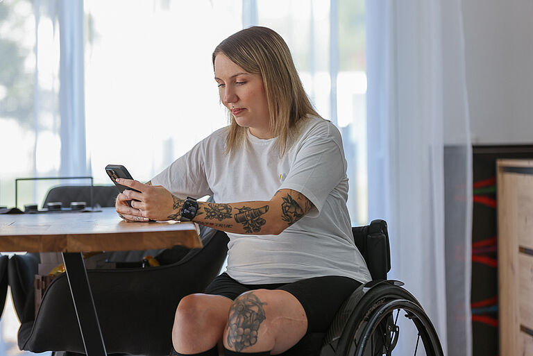 Rollstuhlfahrerin Tamara Fischer sitzt an ihrem Esszimmertisch. Sie schaut auf ihr Handy, das sie in den Händen hält, um ihren Social-Media-Account zu bearbeiten. 