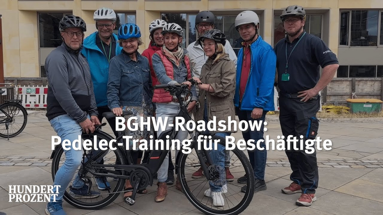 Eine Gruppe von Menschen steht lachend hinter einem Fahrrad. Alle tragen Fahrradhelme, ein Mann sitzt auf dem Gepäckträger. 