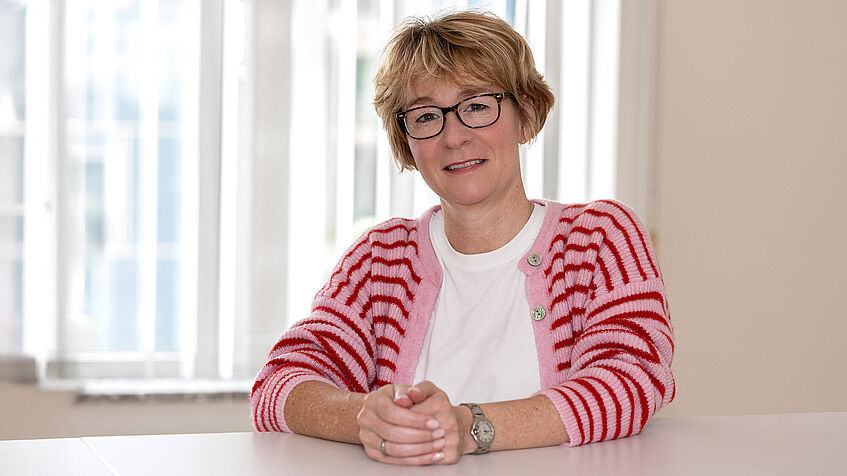 Eine Frau steht an einem Stehtisch. Sie trägt eine Brille und hat eine gestreifte Strickjacke an. Eine Frau steht an einem Stehtisch. Sie trägt eine Brille und hat eine gestreifte Strickjacke an.