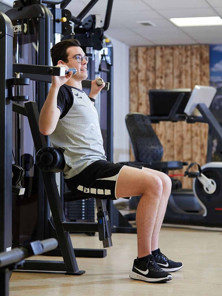 Tim Mayer sitzt in Sportkleidung an einem Kraftgerät und drückt mit zwei Händen Gewichte. 