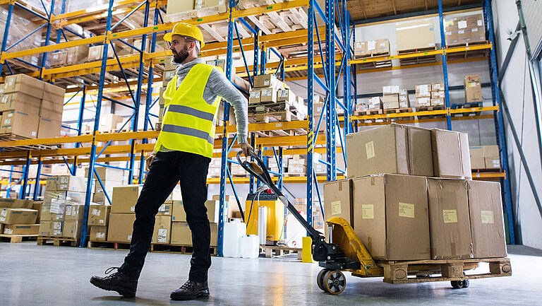 Mitarbeiter im Warenlager zieht eine Mitgängerflurförderzeug mit Kartons hinter sich her.