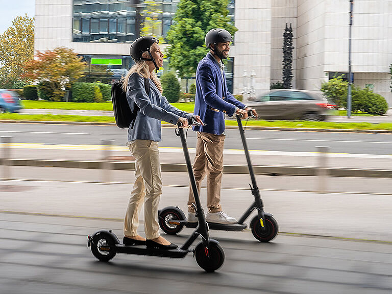 Eine Frau und ein Mann fahren jeweils einen E-Scooter parallel zueinander. 