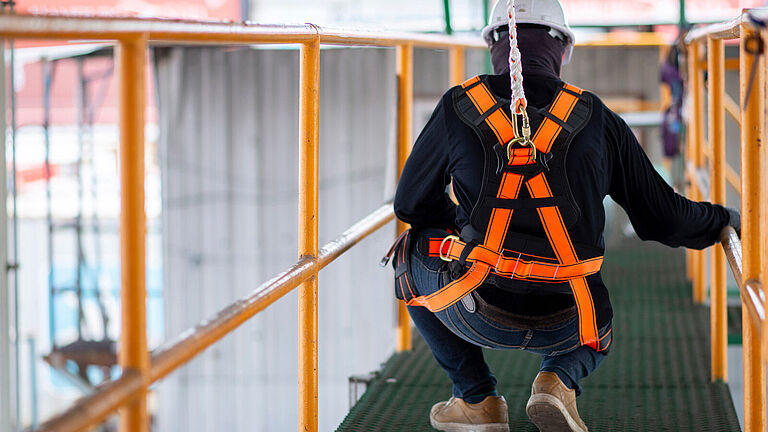 Mitarbeiter von hinten kniet auf einem Gerüst und trägt eine PSAgA mit einer Auffangeinrichtung am Rücken.