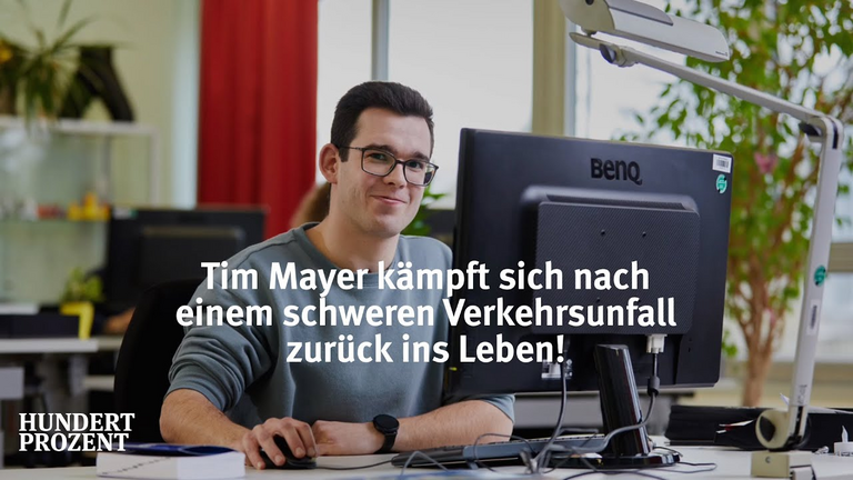 Tim Mayer sitzt hinter seinem Computer-Bildschirm an einem Tisch und schaut in die Kamera. 