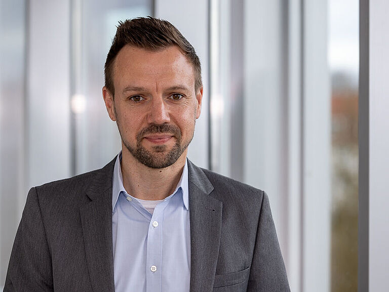 Ein junger Mann mit Bart im grauen Jackett steht vor einem Fensterrahmen und schaut freundlich lächelnd in die Kamera. 