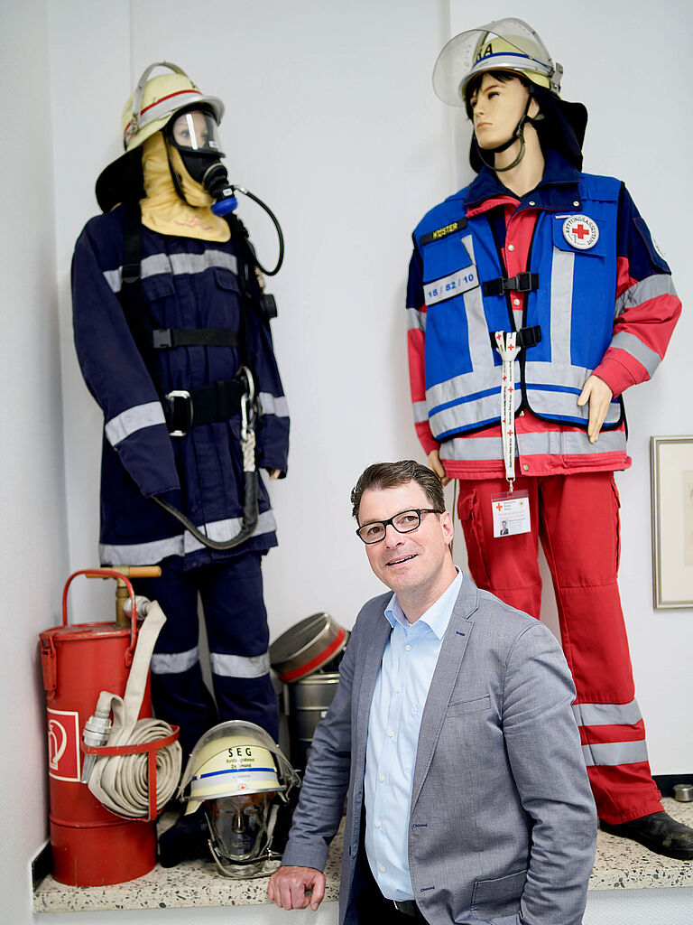 Mann mit Brille steht vor Uniformen eines Feuerwehrmanns und Rettungssanitäter