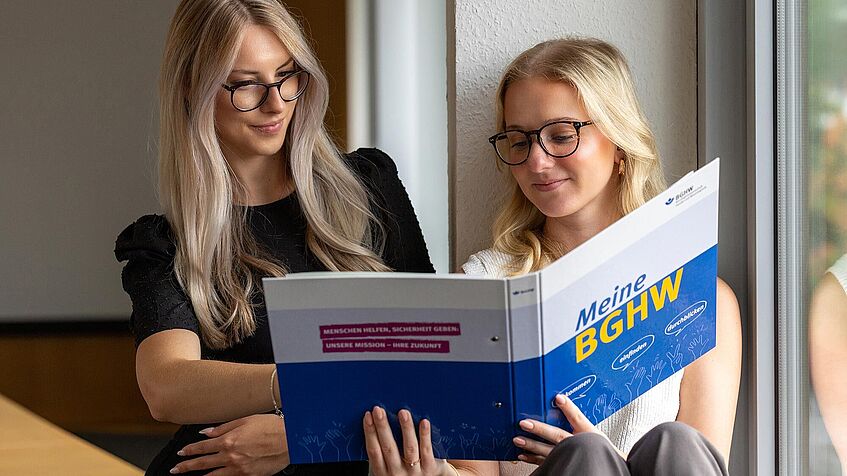 Zwei junge Frauen sitzen am Fenster. Sie studieren gemeinsam eine Informationsmappe.