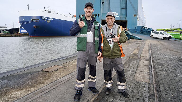 Zwei Hafenarbeiter mit Funkgeräten stehen vor Autotransportschiff im Hafen.