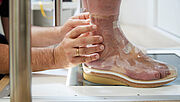 Orthopädie-Schuhmacher Percy Hartmann umfasst mit den Händen zwei Füße in Klarsichthüllen an. Der Mann, dem die Füße gehören, steht mit dem Rücken zu ihm. Diese Folien wurden nach Vorlage der Leisten gefertigt. Bei der sogenannten Folienanprobe wird deutlich, ob die Schuhe passen. 